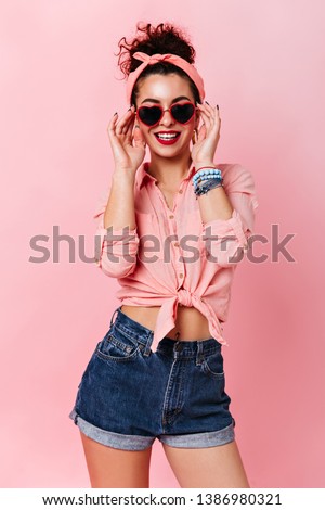 Pin-up girl in headband puts on heart-shaped sunglasses. Portrait of woman in denim shorts and shirt posing on pink background