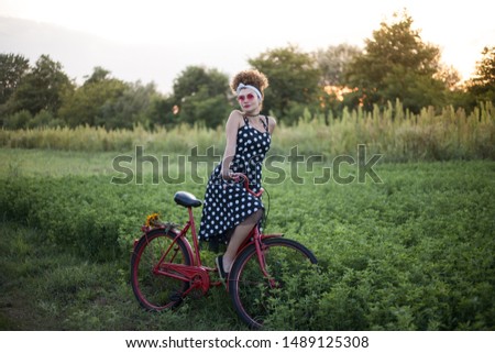 pinup girl with red bike on green field, summer sunset in background