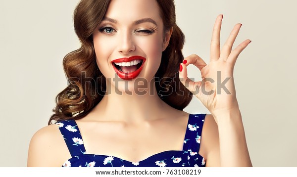 Pin-up retro girl with curly hair  winking,\
smiling and showing OK sign . Presenting your product. Expressive\
facial expressions