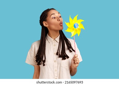 Pinwheel Toy. Portrait Of Woman With Black Dreadlocks Blowing At Paper Windmill, Playing With Hand Mill On Stick, Having Fun, Wearing White Shirt. Indoor Studio Shot Isolated On Blue Background.