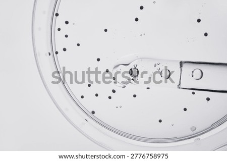 Pipette dropping clear liquid into petri dish with numerous particles, illustrating scientific experiment