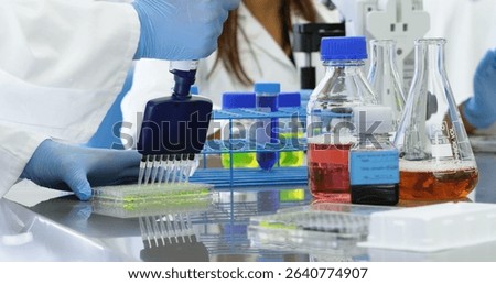 Pipetting gloved hands transferring solution into 96-well microplate at lab bench with tube rack. Scientists, research, precision, innovation, laboratory, collaboration, scientific