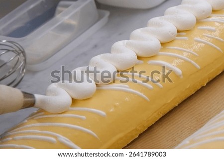 Piping white cream in a zigzag pattern on a yellow roll cake with glossy icing and striped glaze, captured during the final cake decorating process on a marble counter

