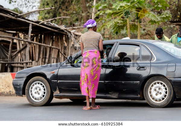 PIRA, BENIN - JAN 12, 2017:\
Unidentified Beninese woman in colored clothes stands near the\
black car. Benin people suffer of poverty due to the bad\
economy.
