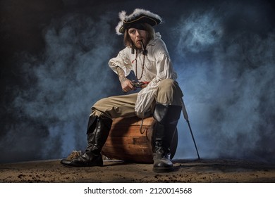 Pirate Filibuster Sea Robber In Suit With Gun And Saber Sits On Barrel. Concept Photo