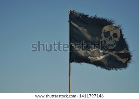 A pirate flag is developing in the sky. The flag is black with a skull. Pirate flag on blue sky background