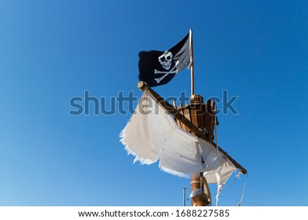 A pirate flag on top of a ship, with a white sail flowing in the wind. The flag is black with a white skull on it. The ship is wooden. There's a clear blue sky in the background. It's a sunny day.