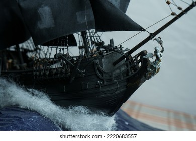 Pirate Ship Model Black Made Of Plywood, With Miniature Figures Of Pirate Sailors, Close-up Details. Bow With Figure Of Woman With Wings. Ship Sails On Waves With Patched Sails