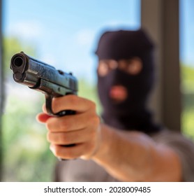 Pistol In Burglar Hand. Man In A Balaclava Aiming With A Gun, Blur Building Interior Background, Closeup View. Intruder, Armed Robber Concept