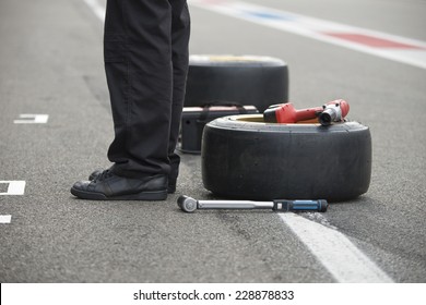 Pit Crew Mechanic Waiting To Get Into Achtion, With Two Replacement Slick Tires A Power Tool And A Torque Wrench Lying On The Asphalt Of The Pit Lane Behind Him