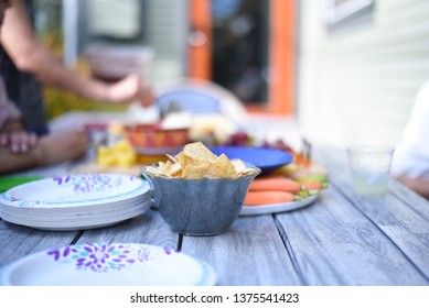 Pita Chips In A Bowl On A Picnic Table Next To Disposable Plates With Assorted Foods Blurry In The Background.