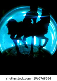 A Pitch Black Shadow Of A Glitter Globe In Carrousel Style Horse And Sat Woman, With Exquisite Beams Of Blue And White Light Piercing Through. The Globe Is Outlined With A Fine Black Shadow.