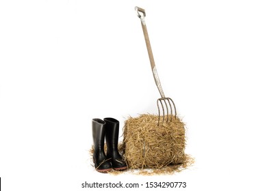 A Pitch Fork Stuck Into A Bale Of Straw With A Pair Of Rubber Bo0ts Isolated On White