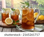 Pitcher of cold iced tea with mint, lemon slices and ice with two glasses ot tea on a wooden table and rural summer background
