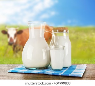 Pitcher, jar and glass of milk on wooden table against cow and blue sky background