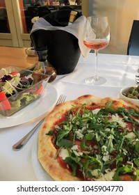 Pizza Al Fresco. An Outdoor Table At An Italian Restaurant Set With Traditional Cuisine Of Brick Oven Pizza, Salad, Olives And Rosé Wine On A White Tablecloth. 