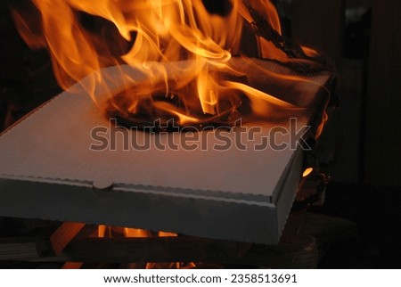 Pizza cardboard box. On the chipped boards folded in a tower lies a white paper box in which there was once a pizza. A fire begins to burn under it, the box also burns with an orange flame.