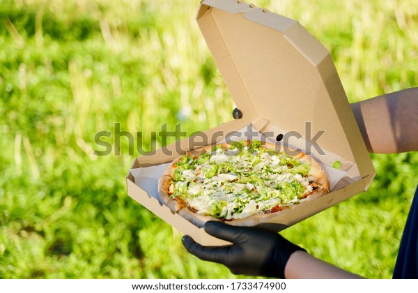 Pizza closeup in hands in medical gloves.\
Courier in medical mask holds pizza in hands and looks to pizza\
with green salad, tomatoes, cheese. Advertise in social network for\
restaurant delivery.