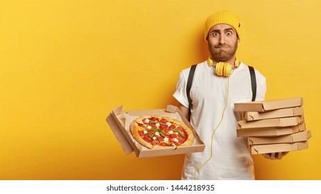 Pizza delivery concept. Handsome courier holds cardboard boxes with pizza, suggests taste delicious fast food, wears hat and t shirt, carries rucksack, poses over yellow background, copy space area