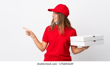 Pizza Delivery Woman Holding A Pizza And Protecting From The Coronavirus With A Mask Over Isolated White Wall Pointing To The Side To Present A Product