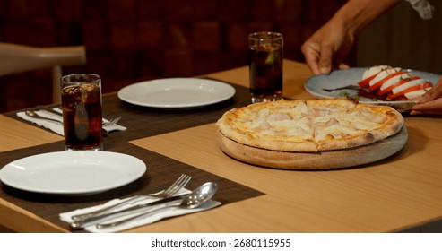 Pizza dinner table setting with drinks, plates, and utensils in restaurant. Person placing Caprese salad. Italian cuisine, dining concept, cozy atmosphere.