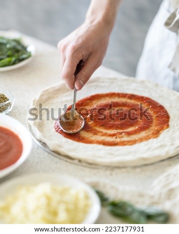 Pizza dough spread with sauce. Process of making pizza in kitchen. Chef spreads red sauce with spoon. Ingredients on table. Light background. View from above. Soft focus. 