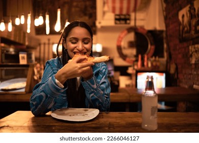 Pizza, food and restaurant where young woman is eating food and enjoying drinks. Hungry female having delicious meal at bar at night. Happy and satisfied foodie having tasty supper at a diner