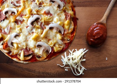 Pizza With Ham, Mushroom And Pineapple Against A Wood Background With Shredded Mozzarella Cheese And Spoon Of Tomato Sauce