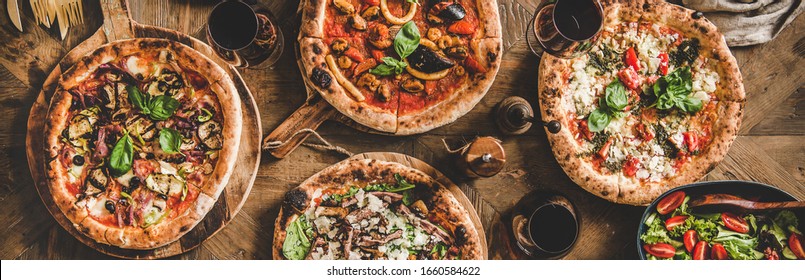 Pizza-Party-Abendessen. Flach gelegene italienische Pizza, Salat und Rotwein in Gläsern auf rustikalem Holztisch, Draufsicht, breite Komposition. Fast Food Mittagessen, Feiern, Sammelkonzept