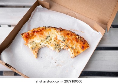 Pizza slice in takeout paper box on a bench. Last bite of pie with teeth marks. Eaten pizza leftovers with crust. Restaurant fast food takeaway or delivery. Delicious Quattro formaggi. Eat outside.