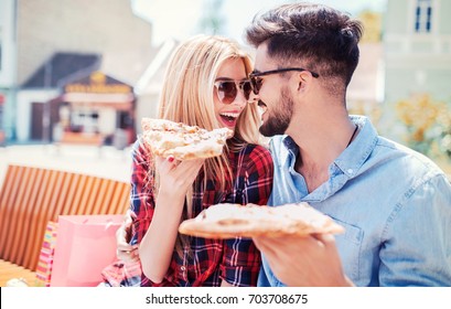Pizza time. Beautiful loving couple sitting on the park bench and eating pizza after shopping. Dating, consumerism, food, lifestyle concept