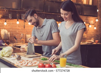 Pizza Time. Young Beautiful Couple In Kitchen. Family Of Two Preparing Food. Couple Making Delicious Pizza. Man Grating Cheese. Nice Loft Interior With Light Bulbs