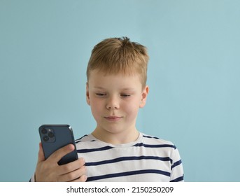 Pkov, Russia - April 06, 2022: A Happy Caucasian Boy Of 9 Years Old, Isolated On A Blue Background, Wearing A Striped Longsleeve Using An IPhone 12 Pro