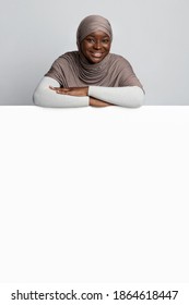 Place For Ad. Smiling Black Woman In Hijab Leaning On Blank White Advertisement Board With Copy Space For Your Design, African Muslim Lady Standing Behind Blank Poster Over Light Studio Background