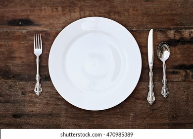 Place Setting Of A Dining Set Of An Empty Plate With Spoon, Fork And Knife Over A Rustic Old Wooden Background. Image Shot From Overhead.