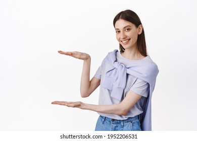 Place your logo here. Smiling cute girl holding empty space between hands, hold box on copy space, showing big size object, standing over white background advertising