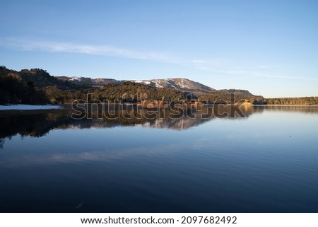 The placid lake at sunset. The clear blue sky, forest and mountains in the horizon, reflected in the water surface. 