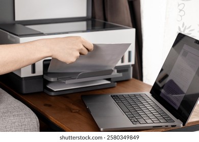 Placing a Document into a Wireless Printer’s Paper Tray, Laptop Open on the Desk for a Home Office Printing Task