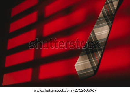Plaid necktie hanging with dramatic shadow lines on vibrant red background creating bold and stylish visual effect.
