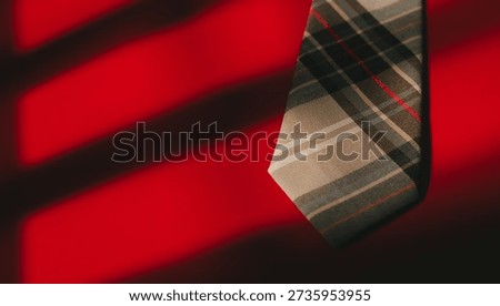 Plaid necktie hanging with dramatic shadow lines on vibrant red background creating bold and stylish contrast