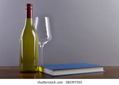 A Plain, Generic White Wine Bottle With A Red Screw-cap Capsule, In Front Of A Light Grey Background On A Brown Wooden Surface, Accompanied By A Wine Glass And Book With A Blue Cover.