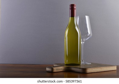 A Plain, Generic White Wine Bottle With A Red Screw-cap Capsule, In Front Of A Light Grey Background On A Brown Cutting Board And Dark Wooden Surface, Accompanied By A Wine Glass.