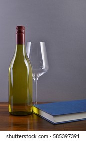 A Plain, Generic White Wine Bottle With A Red Screw-cap Capsule, In Front Of A Light Grey Background On A Brown Wooden Surface, Accompanied By A Wine Glass And Book With A Blue Cover.