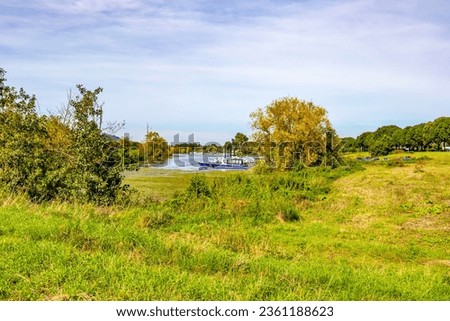Plain with green grass with a small harbor with boat anchored at pier on Maas river in background, green leafy trees, sunny and foggy summer day with blue sky in Eijsden, South Limburg, Netherlands