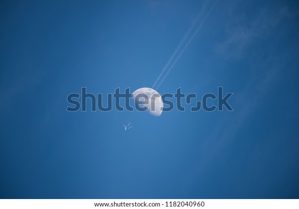 plane passing through the Moon from\
bottom view with long condensation tail on blue\
sky