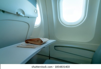 Plane Window. Food In Brown Paper Bag And Plastic Fork And Spoon On Plastic Airplane Tray Table At Seat Back. Economy Class Airplane Window. Inside Of Commercial Airline. Seat With Armchair.