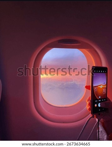 A plane window glows with a soft pink reflection. A hand holds a smartphone close to the glass, capturing the view outside, blending travel, technology, and the beauty of the sky.