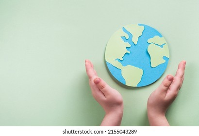 Planet Earth model papercraft in child hands on pastel green background. Environment day, ecology care, sustainable living concept. Top view, flat lay, copy space