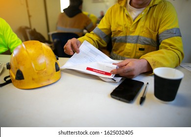 Planner site engineer sitting placing safety hard hat on the table coffee cup pen reviewing JSA risk assessment working at height document prior sign approvals permit by senior site supervisor     
