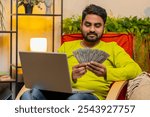 Planning budget. Rich happy Indian man counting money cash use laptop computer calculate domestic bills at home. Hindu guy satisfied of income earnings, saves money for planned vacation, gifts